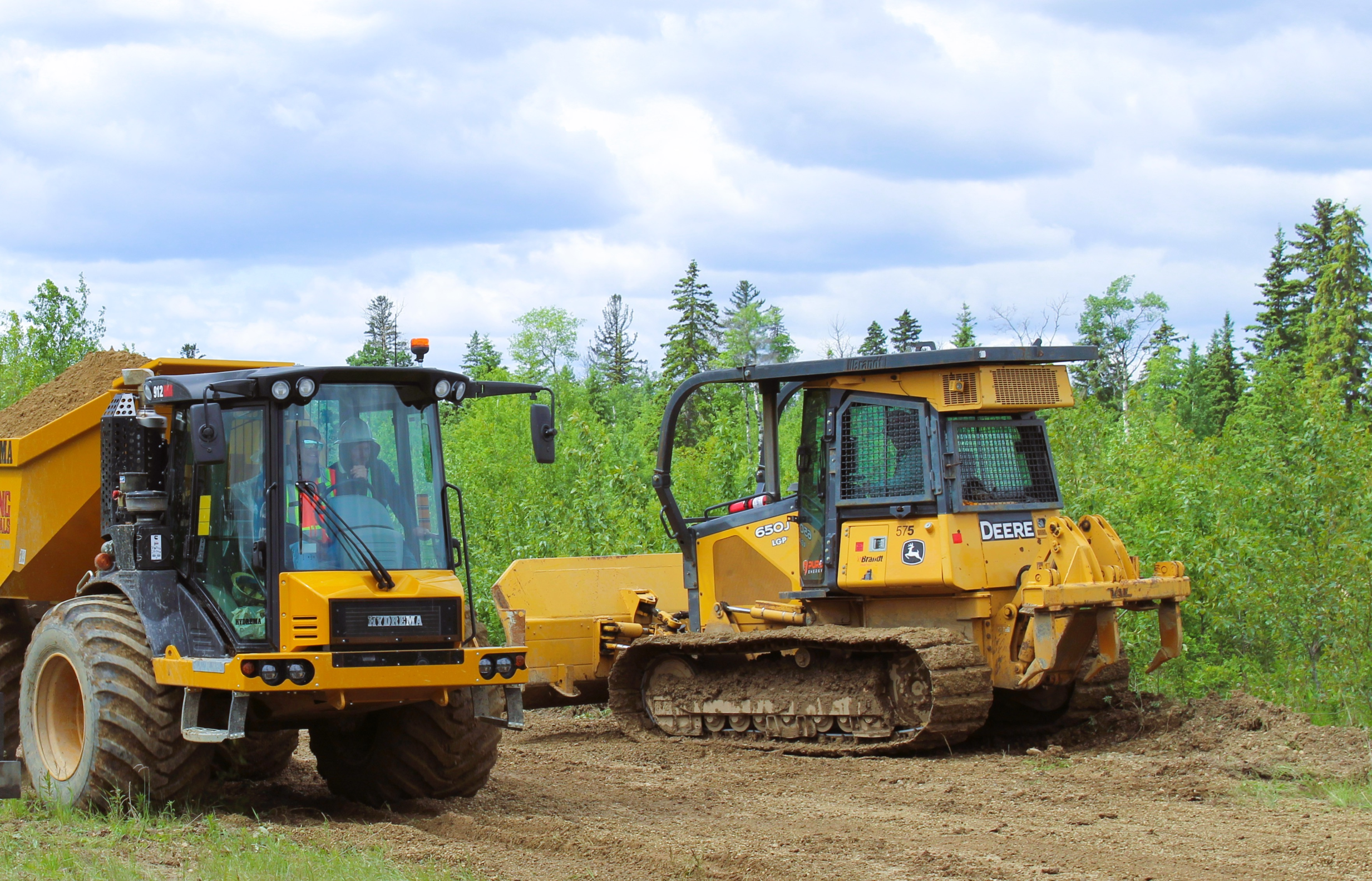 Two heavy construction vehicles working on a dirt site under a cloudy sky.