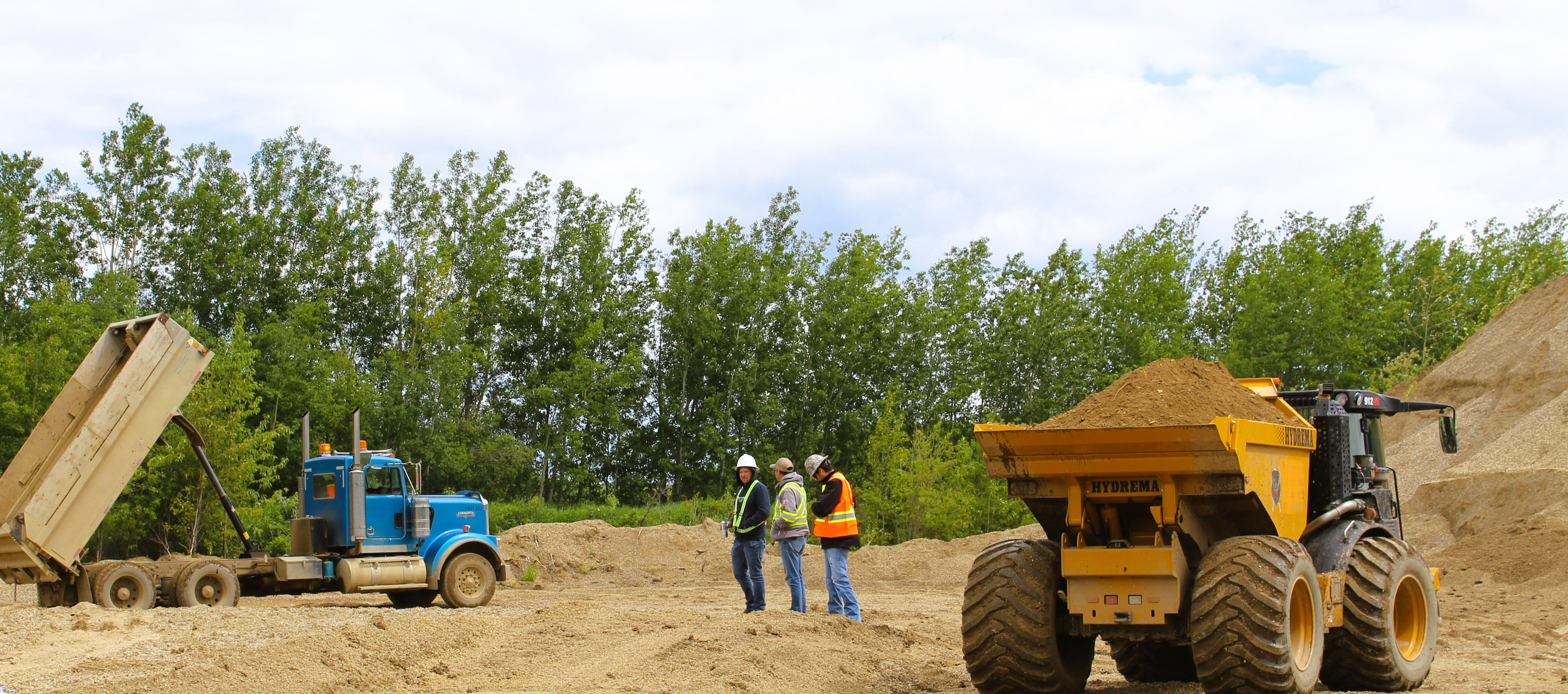 Three construction workers stand on a sandy site near heavy machinery and trees.