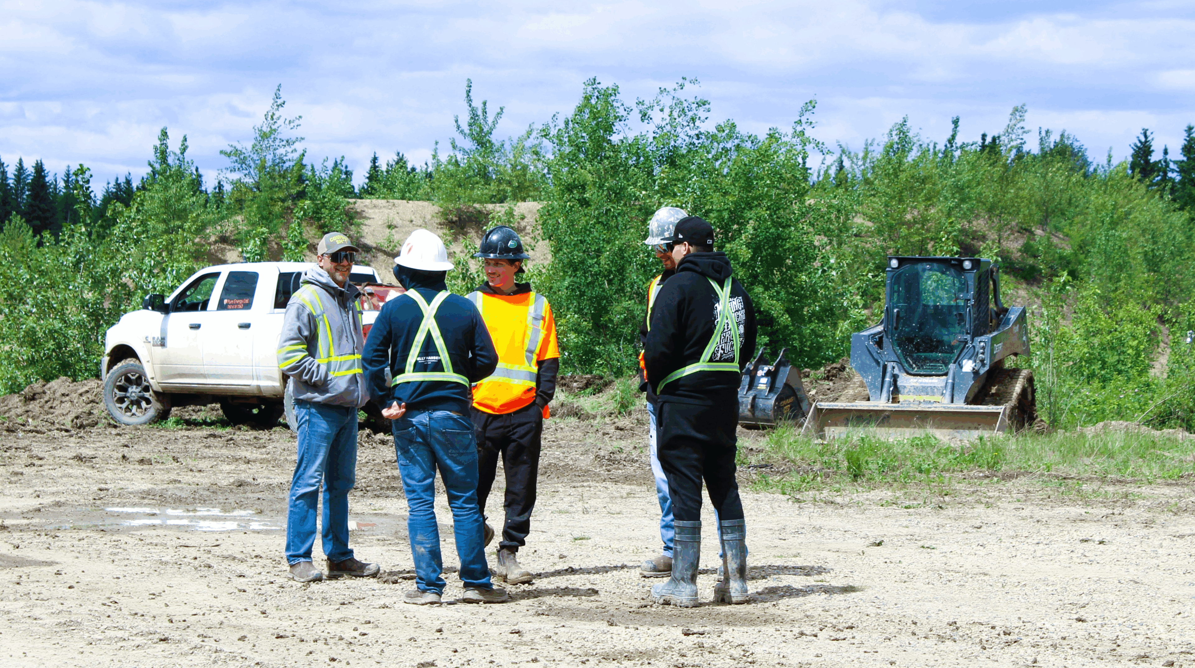 Five construction workers discuss plans on a dirt site with equipment nearby.
