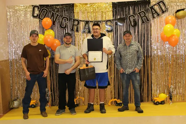 Five men posing at a graduation celebration with decorations and a certificate.